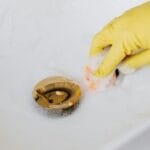 Szybka diagnostyka zapchanego odpływu: co działa, a co szkodzi rurze Ręka w rękawiczce czyści zapieniony, złoty odpływ w zlewie