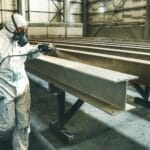 Pracownik w kombinezonie natryskuje metalowe elementy wewnątrz hali